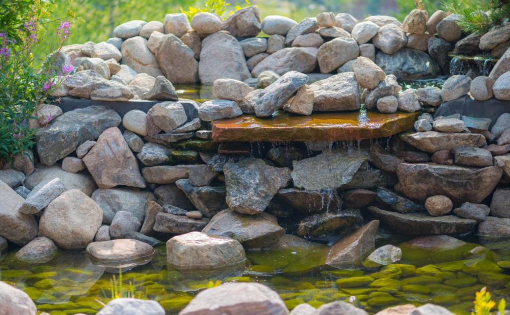 Pequeña cascada de jardín hecha con piedras naturales, rodeada de vegetación y con agua cristalina cayendo sobre un estanque con fondo de rocas