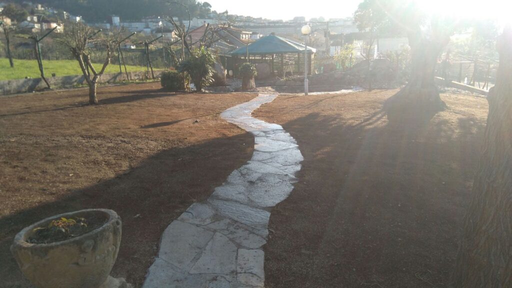 Camino de piedra curvo en un jardín con árboles podados y una glorieta al fondo bajo la luz del sol
